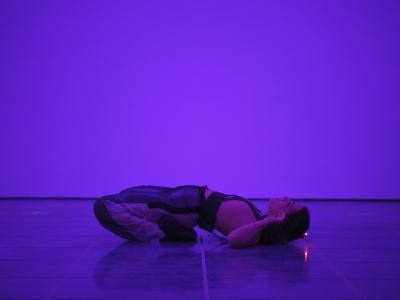 Nour Mobarak reclines with her feet tucked under her buttocks and her shoulders laid flat on the gallery floor. Her elbows are turned upward to bring her open hands towards her ears. A device attached to her head emits an orange light.