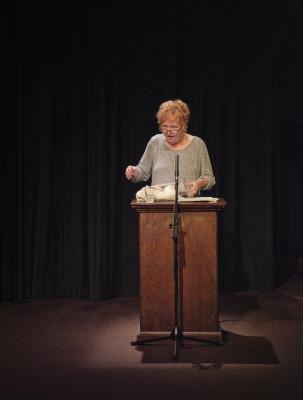 Dorothy Trujillo Lusk speaking at a podium, lit by a spotlight. Backgrounded by black curtains, she wears a grey sweater and glasses. She is in the middle of speech, gesturing with her hands. 