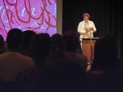 Jordan Abel stands behind a spotlit lectern in the Grand Luxe Hall before a darkened audience. He reads from a book held open in his hands through pink tinted glasses.