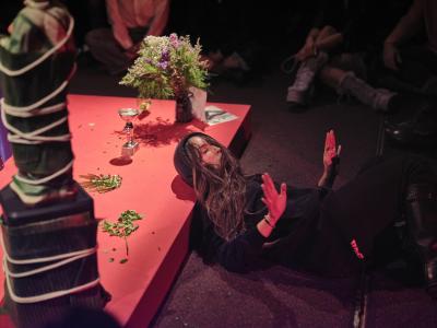 Bhenji’s head and neck rest on the red side of a square platform. Her hair is in movement and her arms are raised from the elbow, her hands in a firm sign of stop.