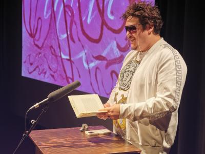 Jordan Abel stands behind a lectern with a book open in their hands. Abel wears pink tinted glasses and a white, gold, and silver outfit fit with rhinestone accents.