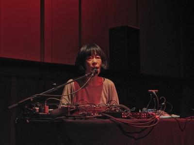 Phew vocalizes into a microphone fixed by a stand to a technology covered table in front of her. She looks straight ahead as a red light contours her left side and diffuses across the panelled wall behind her.