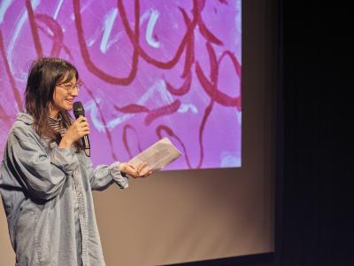 Jacquelyn Zong-Li Ross smiles into a handheld microphone while holding a folded piece of paper. She stands in front of a projection of Jonathan Alfaro’s pink, red, and white monotype print.