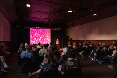 The Grand Luxe Hall is filled with seated audience. A woman in a red dress reading at a wooden lectern in front of a projected pink image.
