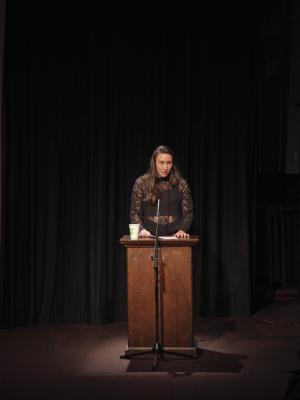 Deanna Fong stands at a wooden lectern in a dark room, backgrounded by black curtains. She is lit by a direct spotlight coming from above. She wears open brown hair and a black see through crochet turtleneck with a black tank top. She is in the middle of speaking, her lips slightly parted — her face appears serious and determined. She pinches a white piece of paper sitting atop the podium. 