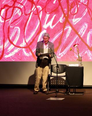 A man wearing a suit stands, reading from a stack of paper. To his right is a black chair and a table covered by a black cloth, topped by a vase with four flowers in it. He is partially illuminated by a pink projected image.