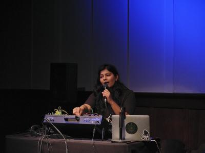 Piu is spotlit from behind a laptop and sound mixer with multicoloured cables in the Grand Luxe Hall. She brings a microphone up to her open mouth and grabs a knob on the mixer with three fingers, looking off into the distance. A diffused blue light is cast onto the panelled white wall behind her.