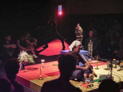 A red spotlight shines on a posed Bhenji. Her legs are spread and bent at the knees, her feet firmly planted on the ground. She is wearing all black and a hood. In the foreground, Tati plays a wooden instrument.