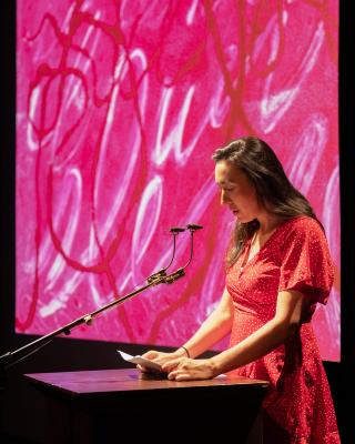 Seen in profile, a woman stands at a wooden lectern reading off a sheet of paper into two small microphones. A pink image decorated with white and red gestural marks is projected behind her.