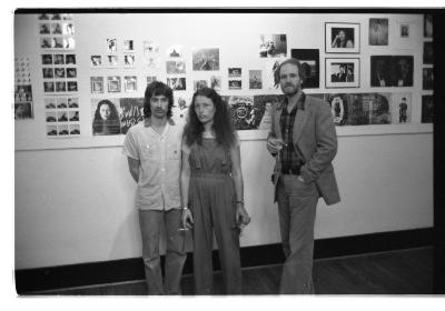 Three people stand in front of a series of exhibited pin-up portraits. Two people stand close together, and on their left, a person holds their hand in their pocket and addresses their attention to the camera. 
