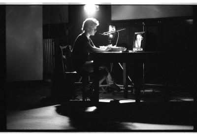 In a wide angle-shot, a person is sitting at the edge of their seat and resting both of their hands on a desk in front of them. Their legs are crossed and they address a television screen that is set toward their face. Above their head, a bright light illuminates their face and casts their body in a soft back-lit glow. A microphone on a stand arcs toward their mouth. 
