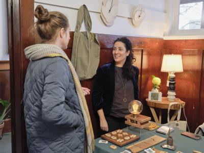 Artist Setareh Yasan listens to a person wearing a quilted blue jacket. The two stand in front of warm toned wood panelling illuminated by a lamp. A table beside them displays a wooden game board, interconnecting wooden slabs, and a wooden structure holding an incandescent light bulb.