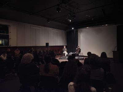 Jayce Salloum and Trey Le sit with microphones in hand while illuminated by spotlights in the back corner of the Grand Luxe Hall. Their bodies face a shadowed audience. Salloum wears a baseball hat shading his eyes and holds his microphone up to speak, resting his other hand open in the air.