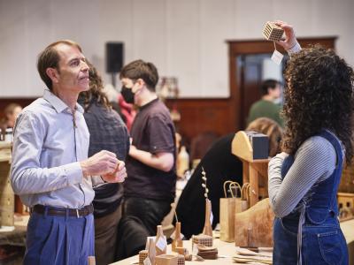 Jody Lentz rests his hands near his waist in anticipation as he looks upwards at a wooden cube held up to the light by a person across from him. Each face of the cube has a five by five pattern of circular holes carved out of its surface, matching the other cubes displayed on the table between them. A tag hangs from the cube that reads AxeWood Inc.