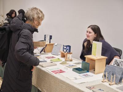 Ruby Lewis from push pull press sits behind a table covered by a white floral tablecloth displaying publications. She looks upwards with a tight smile at a person wearing a long padded jacket standing across the table.