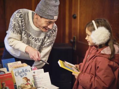 A Toque vendor wearing white and grey sweater and grey toque leans down towards a child with a piece of cardboard in hand. The child wears white fluffy earmuffs and a red jacket while holding a yellow book and a postcard. Beside them are books propped up with text reading C.S. LEWIS THE LION, THE WITCH AND THE WARDROBE, colazione, and Charlottes Web.