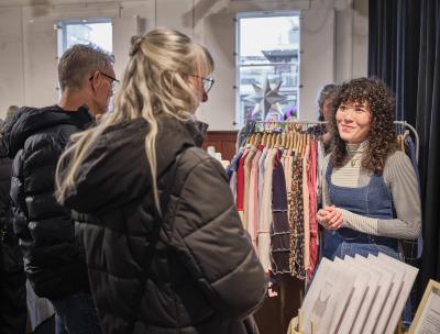 Michelle Larsen from Fortiv stands in front of a full clothing rack with her hands resting together near her waist. She wears a denim top over a striped grey turtleneck. She gazes at two people opposite her with grey hair, both of whom wear glasses and black puffer jackets.