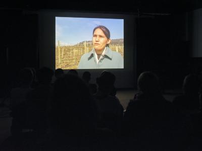 An image is projected against a wall in a dark room. The image is a medium closeup of a person standing in dry farmland on a clear day with rows of wooden posts and distant hills behind them. The warm tones of the image leak out from beyond its frame to reflect against the wooden floor beneath it.