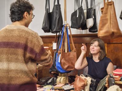 Artist Erin Templeton points with her hand tucked next to her face while looking at a person across from her. Behind her, large leather bags coloured black, brown, and bright blue hang from a wooden stand. Leather pouches and wallets rest in front of her.