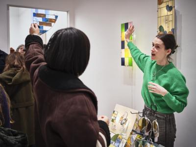 Cora Hall wears a green sweater and fully extends her right arm at an upwards angle. A customer she faces mirrors her gesture, holding a piece of stained glass in the shape of a quarter circle up to the light.