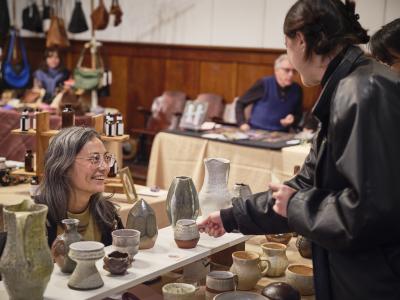 Kristine Aguilar looks up with a wide smile from behind a display of ceramics towards a customer wearing a black leather jacket. The customer grabs the knob handle of a small ceramic pitcher with one hand.