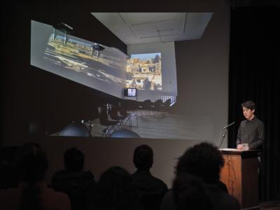 Trey Le is spotlit from behind a lectern in the Grand Luxe Hall. He stands in front of a projected image of a new media installation in a gallery space that features asymmetrical wall projections, a CRT monitor, and scattered chairs.