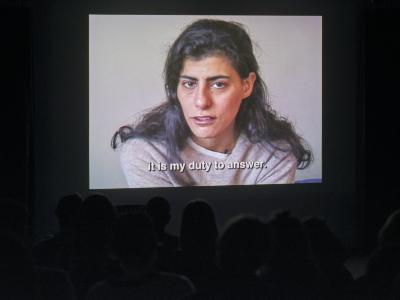 An image is projected against a wall in a dark room, making the outlines of the audience’s heads barely visible. The image is a medium close up of a person in front of a white wall staring straight ahead. A caption underneath the person reads it is my duty to answer.