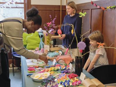 A child sits with their hands on their knees behind a table displaying brightly coloured fabric dolls, bows, and beaded keychains. A customer leaning down towards the table grabs the ring of a keychain with a grin. Artist Justine Leavers is to their side looking downwards while holding a brown object resembling a cardboard container.