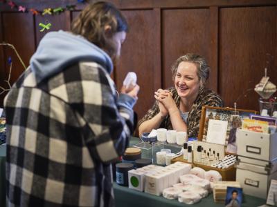 A Toque vendor smiles from behind a table displaying candles, perfume bottles, and other packaged objects. The vendor sits with their hands interlocked and elbows resting on the tabletop looking at a customer bringing a round paper covered object up to their face.