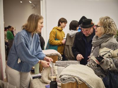 A Toque vendor purses their lips and makes eye contact with a customer they face. The vendor wears a blue linen shirt and gently pinches a piece of fabric lying on the table situated between the two people. To their side, another person studies the tabletop.