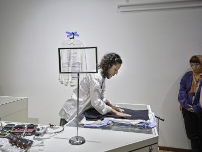 Katayoon Yousefbigloo flattens a pile of blue and white T-shirts against a glass countertop with her hands. The counter is C-shaped and its other side is covered by guitar pedals and a metal signage stand. The top of the metal stand is an empty rectangle that frames part of the white dress shirt hanging behind the counter against the white wall of the gallery.
