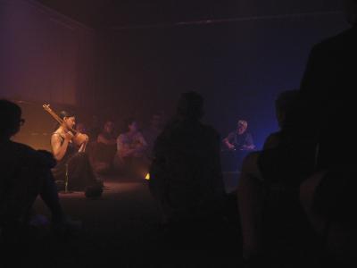 Samita Sinha and Ash Fure are framed by the bodies of audience members seated alongside them on the floor of the Grand Luxe Hall. Sinha holds an ektara beneath the warm glow of orange light while Fure is enveloped in a purple haze, lightened partially from the blue light to their side. Fure has both hands on a control board.