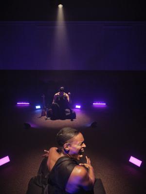  Okwui Okpokwasili and Samita Sinha are independently spotlit while seated in front of one another in the Grand Luxe Hall. Okpokwasili turns her head sharply to the left while tensing her face as if yelling. Sinha drops her head low while raising the bunched fingers of one of her hands. Blue and purple rectangular lights scatter the floor, diffusing their pigment throughout the space.