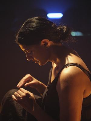Samita Sinha rests her eyes closed with her hands gesturing in a pinching motion near her chest. She is spotlit by warm lighting while a shadow is cast across her face, leaving only the edge of her face and tip of her ear highlighted in orange. An oblong blue light is in soft focus above her head.