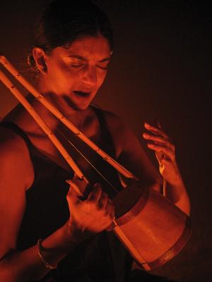 Samita Sinha scrunches her eyes closed while vocalizing. She grips an ektara with a tight fist, leaving her other hand gently suspended in air. Saturated orange-red lights the space, turning all shadows into a near black.