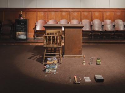 Two wooden plinths background a wooden chair, a stack of books covered with feathers, and two neat rows of objects on the ground. The objects consist of an olive jar, a glass filled with metal tools, a handbell, a cassette player, two cassettes, a letter opener, a hammer, and a knife.