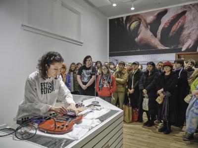 Katayoon Yousefbigloo leans over a red electric guitar resting on a glass countertop. She holds a smartphone displaying an illustration of an eye on top of the guitar strings with one hand and points her index finger at the smartphone screen with another. An onlooking crowd stands to her side in the gallery space. Behind the crowd is an image-covered wall depicting an extreme close-up of a person wearing gold eyeshadow and glossy red lipstick.