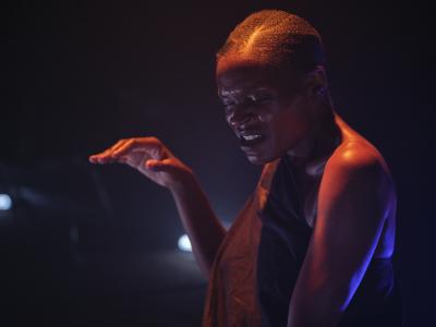 Okwui Okpokwasili furrows her brows while vocalizing in a dark room with round white lights lying on the ground. Her tense expression is contrasted by her hand gently resting in the air with its palm facing downward. An orange light warms the top of her head, shoulder, and fingertips while a blue light emanates from behind, cooling the hue of the background and leaving a bright highlight on her back.