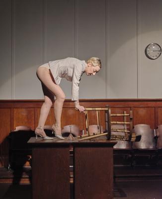 Marie Segolene Brault grabs the legs of a wooden chair lying on its back. Both Brault and the chair are on an elevated wooden platform with an upside down martini glass positioned between them.