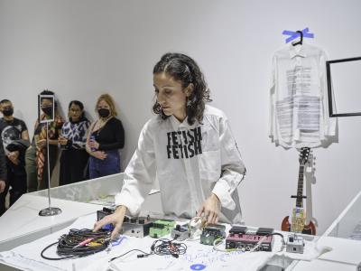 Katayoon Yousefbigloo reaches over a glass counter covered by guitar pedals to grab a pile of TS cables. She wears a white dress shirt with text reading FETISH FETISH printed in black near its collar. Another white dress shirt with a large block of black text printed on its front is hung on a black clothes hanger and affixed to the white wall behind her via two pieces of blue painters tape.