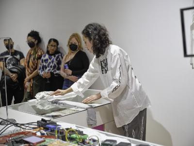 Katayoon Yousefbigloo folds a grey T-shirt widthwise into a long, narrow shape on top of a glass counter, leaving the bottom of the shirt draping off the counters edge. Although partially obstructed by the folds in the fabric, vertical text printed on the arm sleeve and pant leg of Yousefbigloos outfit reads AUTONOMY, AUTO, AUT, and AUTONOM. Four people lean against a wall to her side with gazes fixed on the glass countertop.
