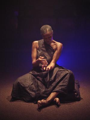 Okwui Okpokwasili sits cross-legged under a warm spotlight with a blue light emanating from behind her back. Her head faces downwards at her arms outstretched in front of her, gesturing as if pouring something from one hand into another. Her arms are foregrounded by her halter strap dress made of thin black linen.