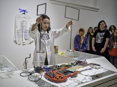 Katayoon Yousefbigloo holds two metal signage stands over a red electric guitar connected to a row of guitar pedals. A smartphone rests on the guitars base and a lottery scratch card rests on its strings. The signage stands have round, reflective bases that attract the gaze of the crowd to her side and Yousefbigloo herself.