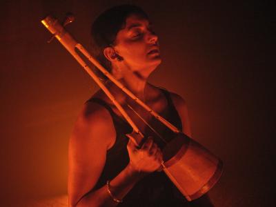 Samita Sinha relaxedly faces upwards with her eyes closed. She holds an ektara in an arm tucked tightly to her side, forearm adorned by a single mental bracelet. Saturated orange-red light emanates from below, filling the space with a warm hue and dark shadows.
