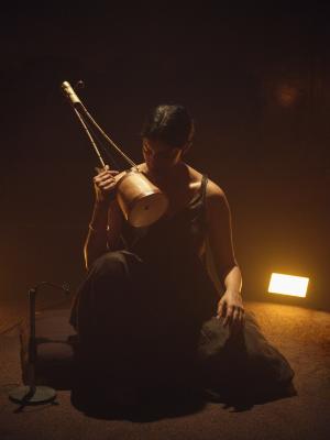 Samita Sinha sits atop a cushion on the floor with one knee raised. She raises the base of the ektara up to her downward gazing face, resting the arm holding the instrument on her raised knee. The edges of her hair, fingers, neck, and collarbone are all defined by the glow of a warm spotlight and a rectangular orange light on the floor behind her.