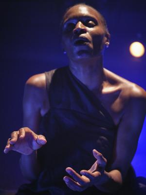 Okwui Okpokwasili is in soft focus with her eyes closed and lips loosely pursed. Her elbows are tucked to her side while her hands are extended outwards into clear focus, one with its palm facing upwards and the other facing downwards. A bright white light shines down onto her from above while saturated blue lights both her underside and the background.