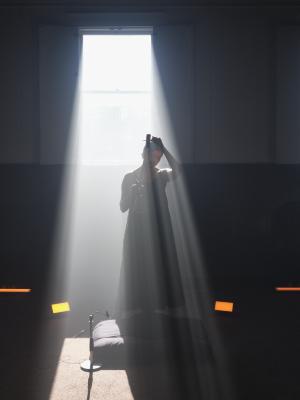 Samita Sinha stands beneath bright white light entering a dark room from a tall window frame only shadowed by the outline of her body and the ektara raised in her hands. A quilted cushion and rectangular orange lights rest on the carpeted floor beneath her feet. The subsurface scattering of light highlights the tip of her thumb and apex of her ear.