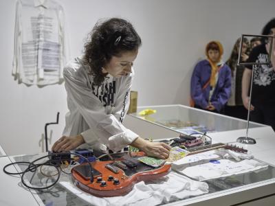 Katayoon Yousefbigloo crosses her hands over a red electric guitar and a row of connected guitar pedals resting on a glass counter. She adjusts a knob on the pedal nearest the guitars base while scratching a lottery scratch card that rests on the guitar strings.