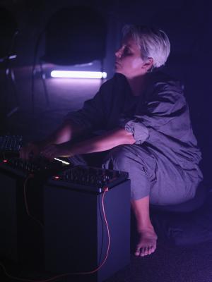 Ash Fure sits barefoot atop cushions in a crouched position behind three elevated sound mixers, adjusting dials from the centre board with both hands while gazing outwards. Soft purple light illuminates half her body and the underside of an empty chair in the background. The rest of the space falls into a cool indigo shadow.