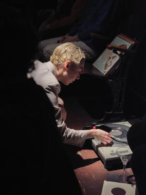 Marie Segolene Brault kneels down adjusting the arm of a record player resting on the ground. A stack of records rests on a nearby chair.